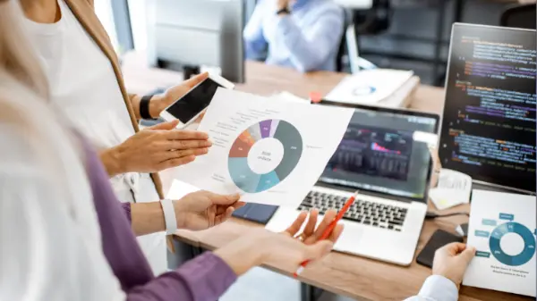 Plusieurs mains manipulent des graphiques circulaires devant un ordinateur affichant du code.