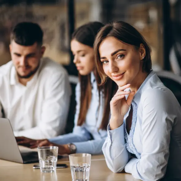 Jeune femme souriante au bureau, main sous le menton, avec des collègues travaillant derrière elle.